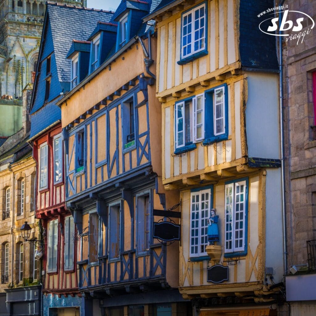 Edifici colorati con intelaiatura in legno e finestre multiple fiancheggiano una strada stretta come una Bozza automatica. L'architettura presenta un mix vibrante di tonalità gialle e blu, adornate da dettagli decorativi e un cielo luminoso sopra.