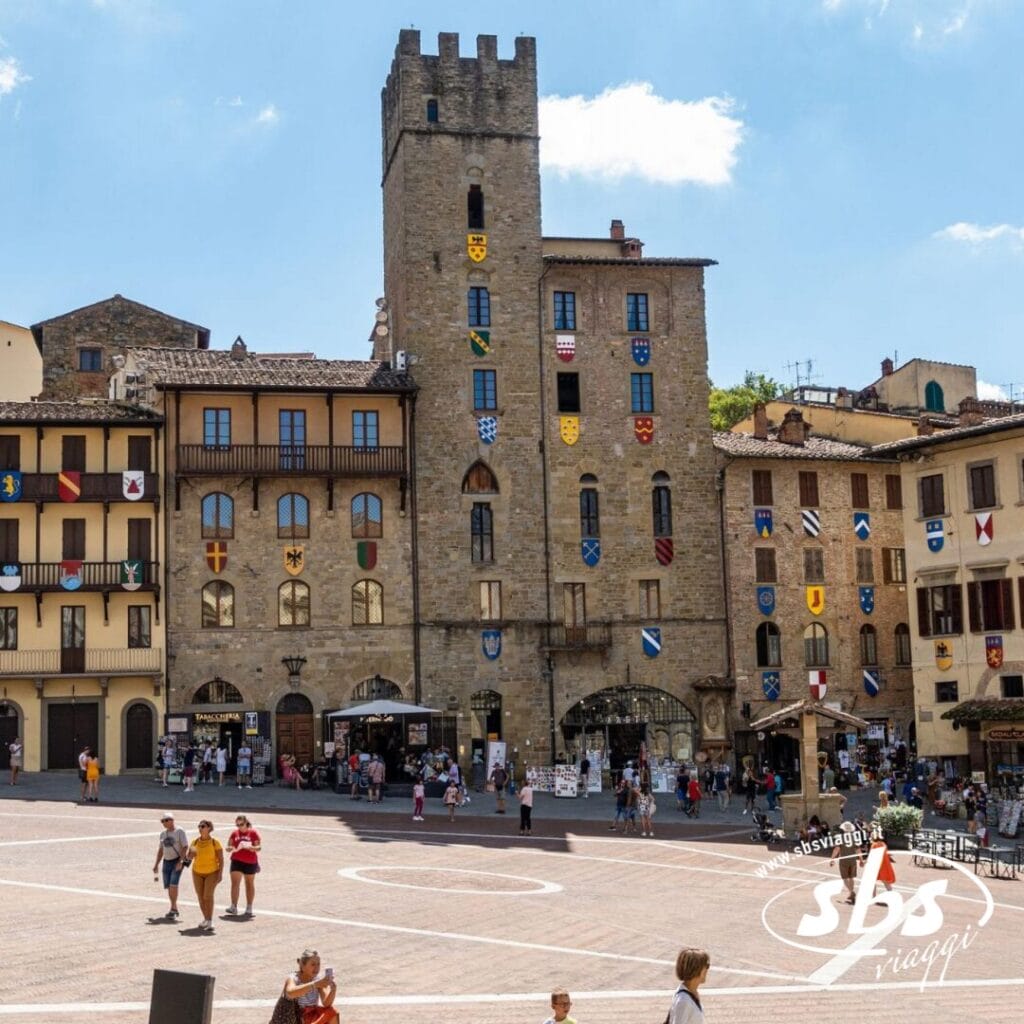 Antichi edifici in pietra con stemmi fiancheggiano una piazza in una città europea, che ricorda Siena. Una folla di persone passeggia attraverso la zona panoramica, incorniciata dal cielo luminoso ed evocando il fascino senza tempo della Val d'Orcia.