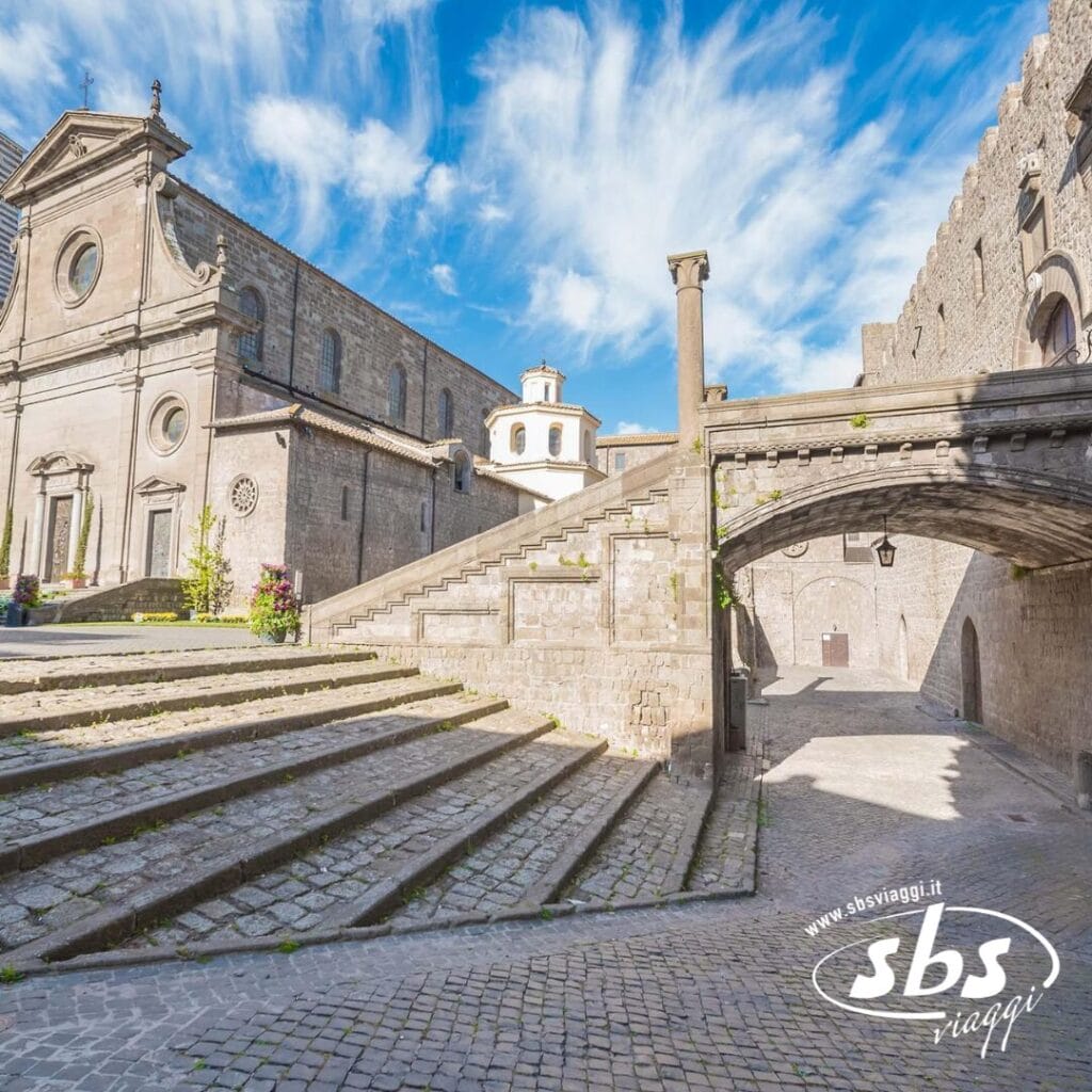 Edifici storici in pietra con gradini e un arco sotto un cielo azzurro brillante, caratterizzati da dettagli in pietra lavorata e sentieri acciottolati, vi invitano a esplorare il fascino dell'Etrusco durante il vostro Tour della Tuscia.