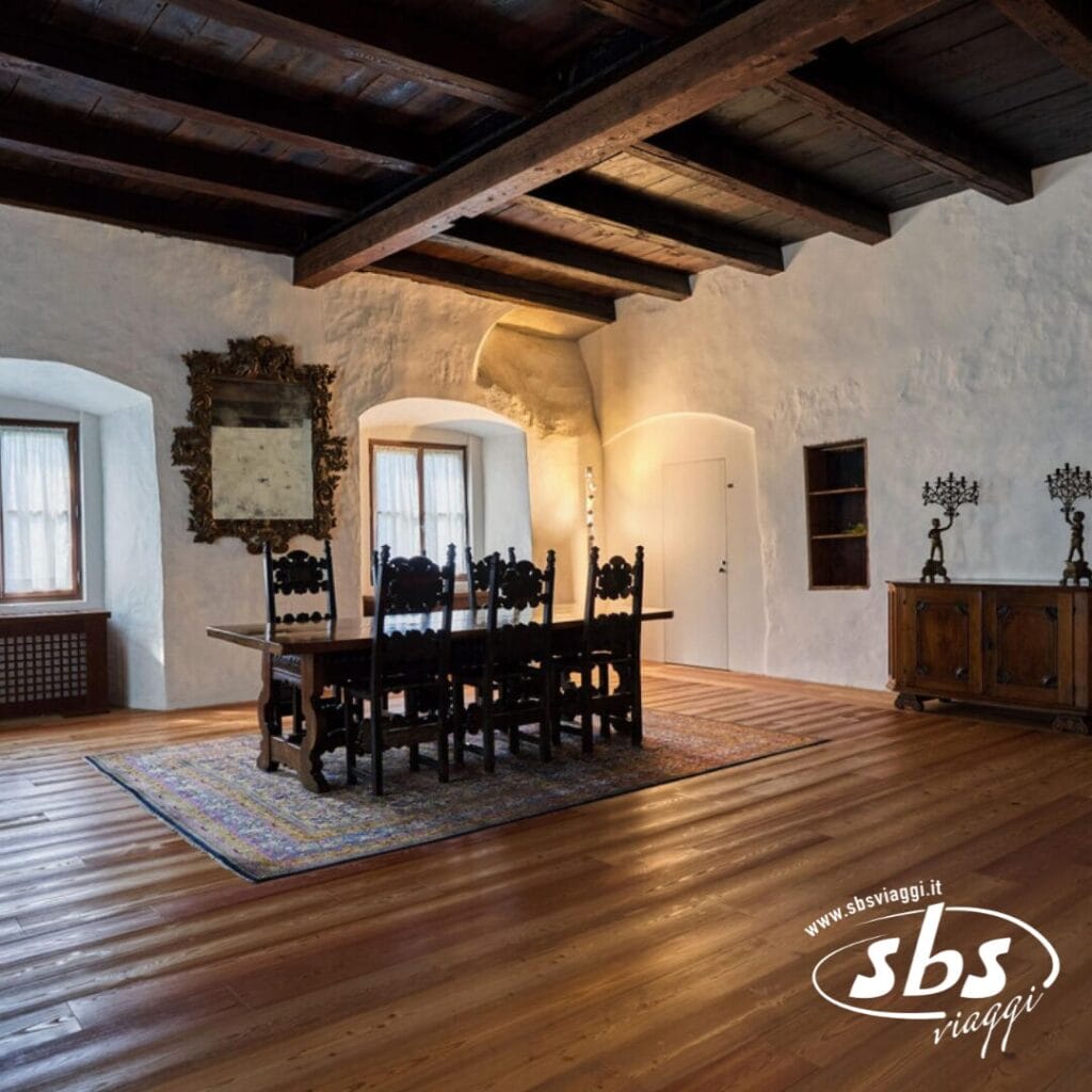 Una sala da pranzo con tavolo e sedie in legno, specchio decorato e travi in legno sul soffitto trasuda fascino. Il pavimento in moquette contrasta elegantemente con le pareti bianche e le porte ad arco. Un logo accenna all'ispirazione tratta dal fascino senza tempo dei Castelli del Trentino.