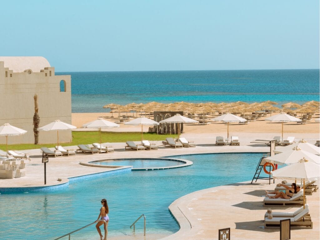 Nel lussuoso Sunrise Anjum Resort, una donna scende con grazia i gradini della piscina tra lettini e ombrelloni. Il mare mozzafiato e la spiaggia sabbiosa creano uno sfondo pittoresco sotto il cielo azzurro e terso, incarnando l'essenza di una fuga a 5 stelle.