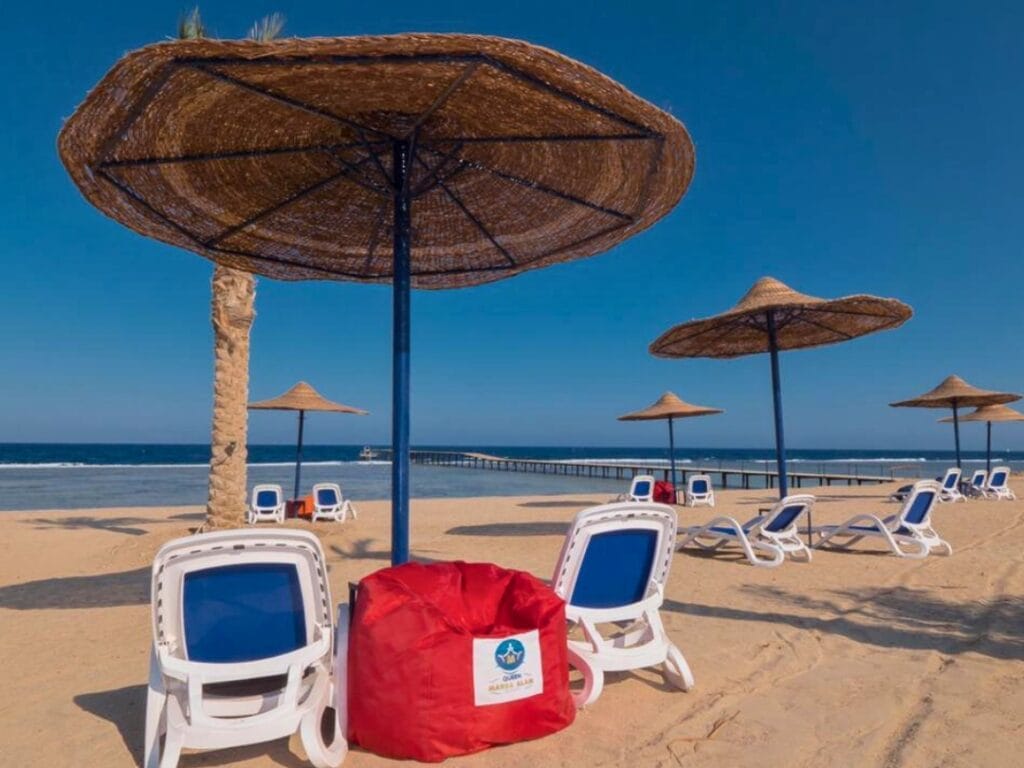 Una tranquilla scena di spiaggia con ombrelloni di paglia e sedie a sdraio bianche è esaltata da un vivace pouf rosso appollaiato sulla riva sabbiosa. Nelle vicinanze, un molo si estende nel mare sotto il cielo azzurro e limpido, offrendo una fuga idilliaca in questa bozza automatica di paradiso.