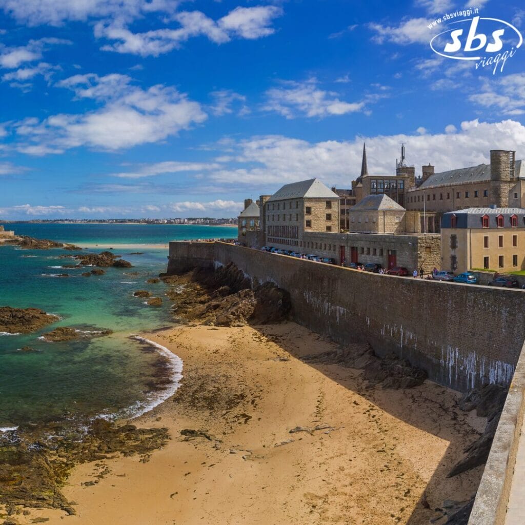 Il panorama costiero presenta una spiaggia sabbiosa, una costa rocciosa ed edifici storici sotto un cielo azzurro brillante con nuvole: una scena idilliaca che ricorda una bozza automatica che cattura la perfezione della natura.