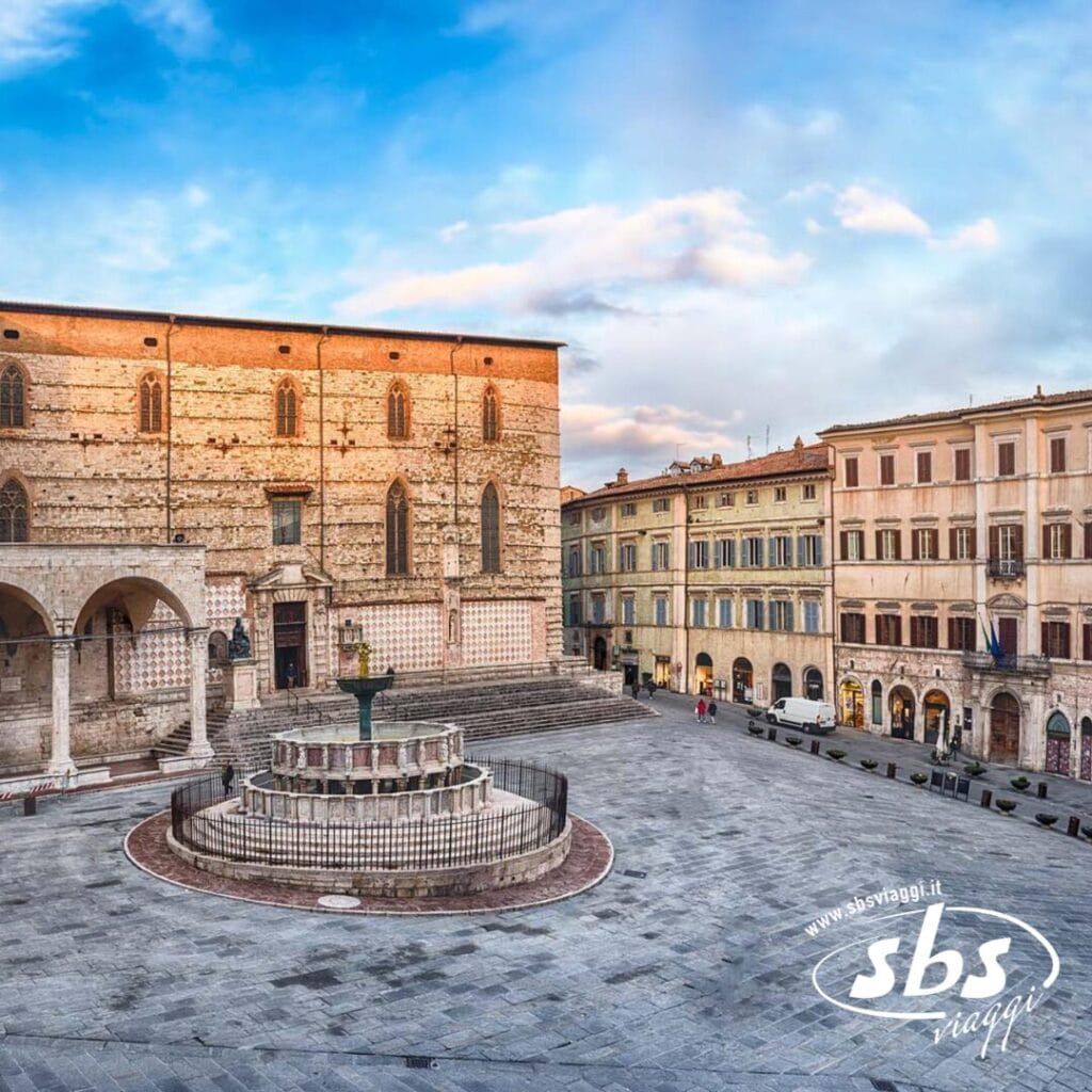 Piazza storica con fontana centrale, circondata da vecchi edifici sotto un cielo azzurro. Il terreno lastricato e l'architettura in pietra riecheggiano il fascino dell'Umbria medievale. Il logo