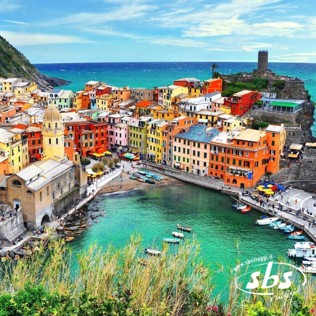 Edifici colorati circondano un porto a Vernazza, Italia, parte delle meravigliose Cinque Terre. Le barche punteggiano l'acqua sullo sfondo di una costa frastagliata e cieli parzialmente nuvolosi, catturando l'essenza di questa pittoresca regione perfetta per chi è in viaggio con il Tour Toscana.