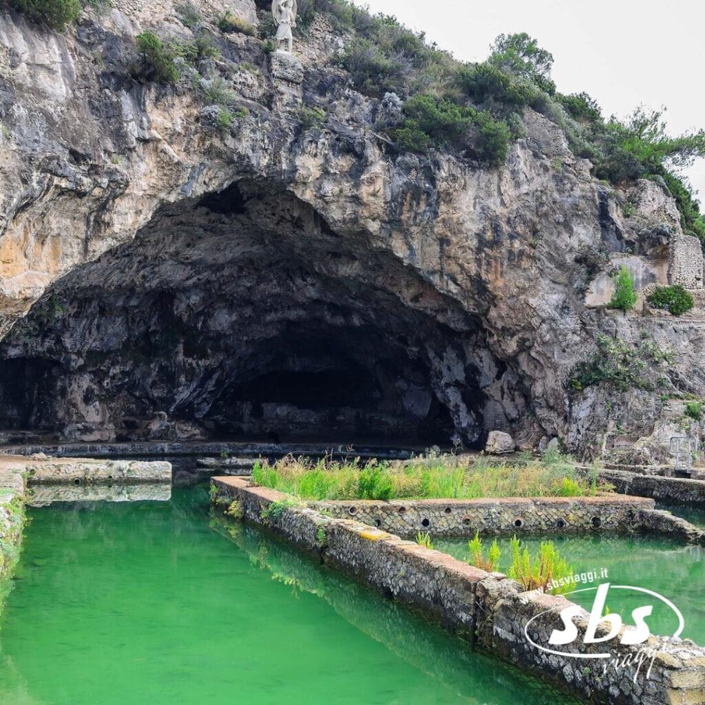 Un ingresso di grotta naturale con acqua verde si apre davanti a voi, delimitato da piattaforme di pietra in mezzo al terreno roccioso e alla vegetazione lussureggiante della Riviera di Ulisse. Un logo adorna l'angolo in basso a destra.
