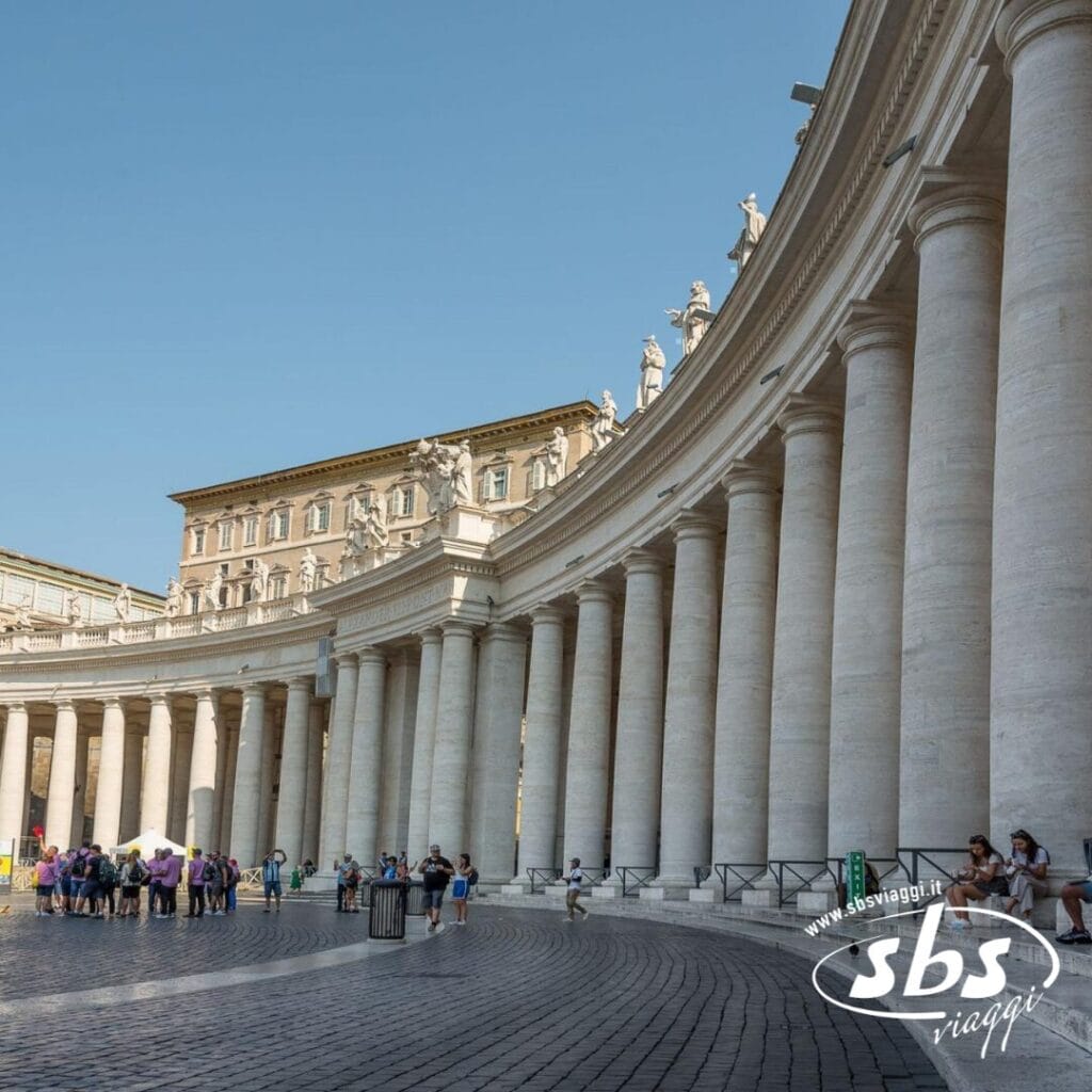 Nel cuore della Città Eterna, il colonnato curvilineo di Piazza San Pietro, Città del Vaticano, accoglie i turisti che passeggiano e si riuniscono sotto un limpido cielo azzurro durante il loro tour di Roma.