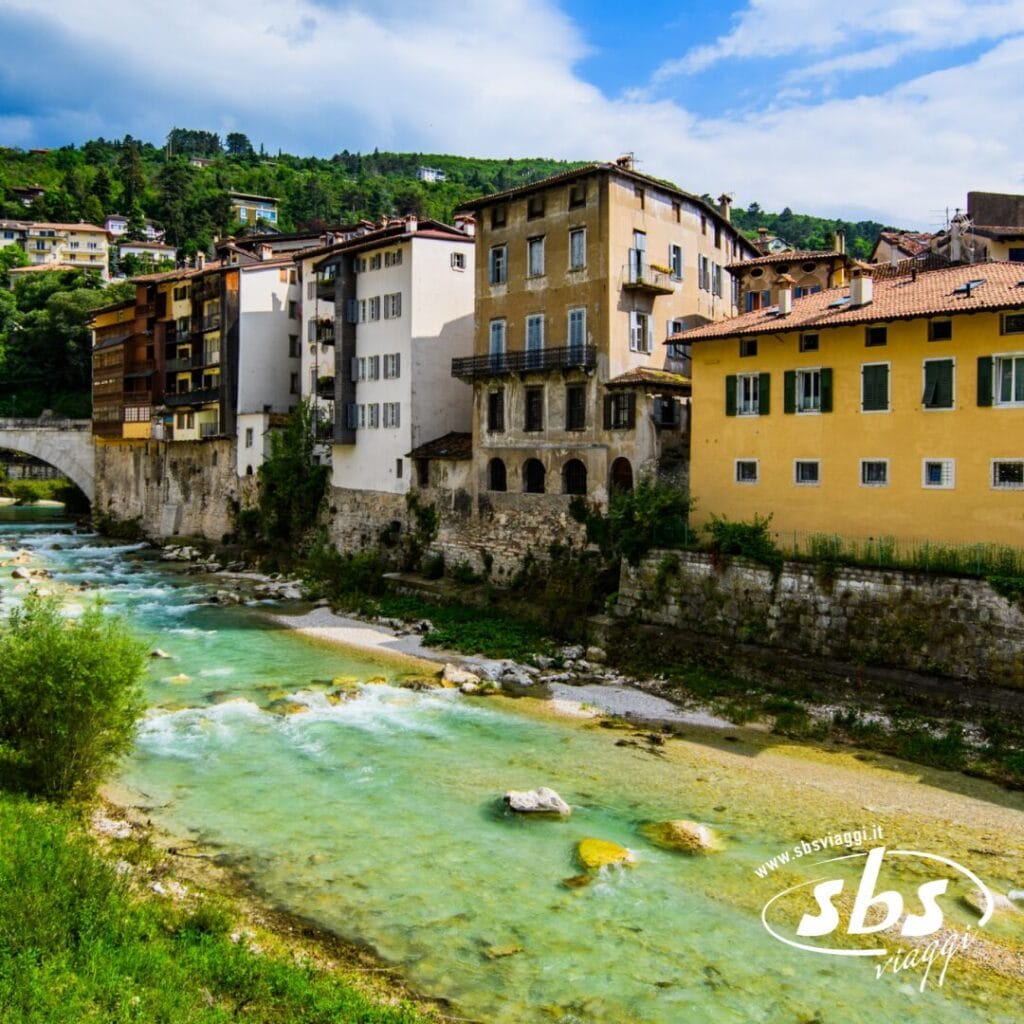 Edifici storici costeggiano la riva di un fiume in una città italiana, con un ponte di pietra sullo sfondo e un cielo limpido sopra: la cornice perfetta per un tour tra i gioielli architettonici del Trentino Alto Adige.