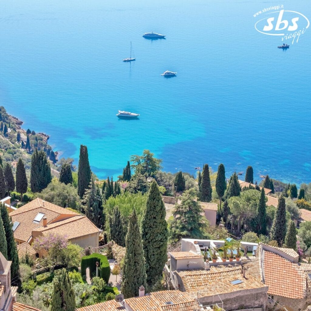 Dalla prospettiva di Bozza automatica, la scena si dispiega: una vista dall'alto di un villaggio costiero con case con tetti in terracotta immerse in una vegetazione lussureggiante, il tutto rivolto verso un mare azzurro e calmo punteggiato da diversi yacht.