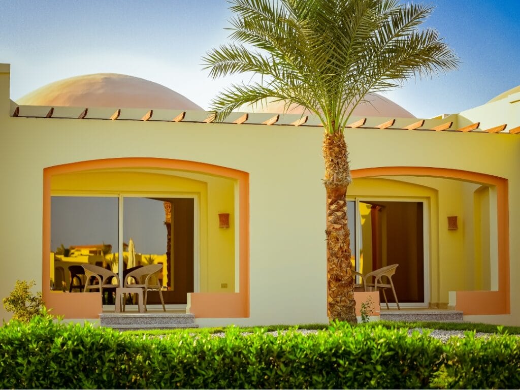 Edificio giallo con due aree salotto esterne a specchio separate da una palma centrale. Ogni area, come una bozza automatica dell'altra, presenta porte scorrevoli in vetro, sedie e tavoli incorniciati da archi. È graziosamente fronteggiato da una rigogliosa siepe verde.