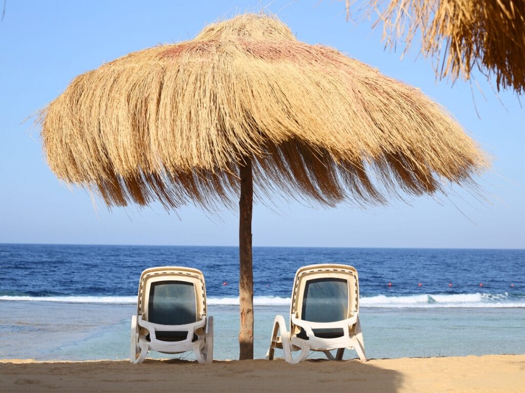 Due sedie a sdraio vuote sono sistemate sotto un grande ombrellone di paglia su una spiaggia sabbiosa di fronte all'oceano, creando la perfetta bozza automatica per una fuga serena.