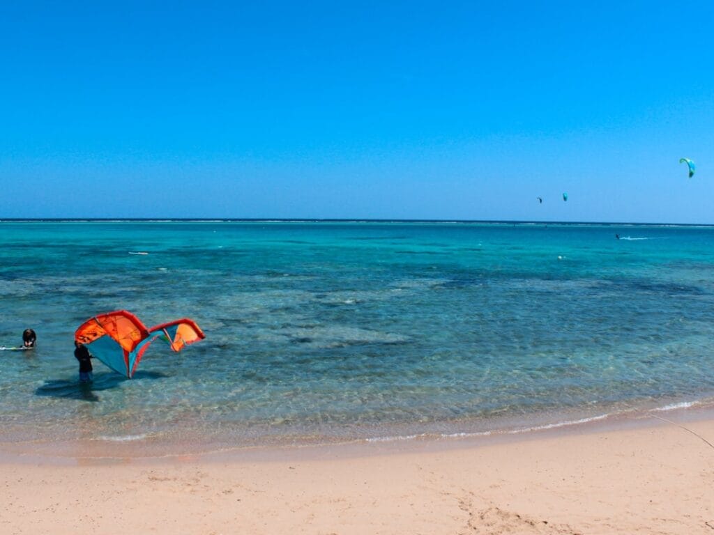 Una persona con l'attrezzatura per l'aquilone è in piedi su una spiaggia sabbiosa, circondata da acque turchesi sotto un cielo azzurro e limpido, come se fosse a bordo di una bozza automatica e stesse catturando la perfezione della natura.