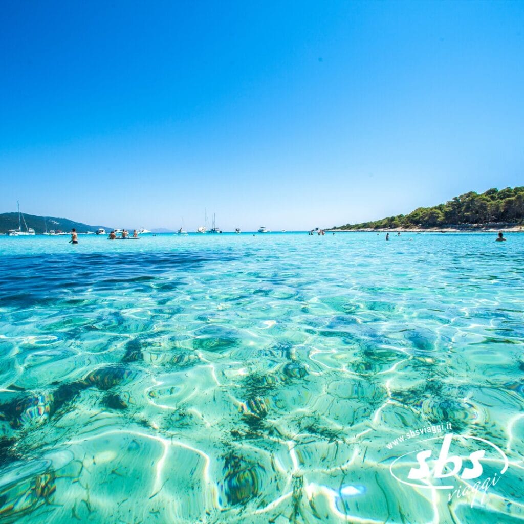 Sotto un cielo azzurro brillante, il mare turchese e limpido ospita alcuni bagnanti, con barche che punteggiano l'orizzonte e verdi colline che incorniciano la scena. Perfetto per una Vacanza Evento under 28 a Pag, dove l'energia vibrante incontra paesaggi sereni.