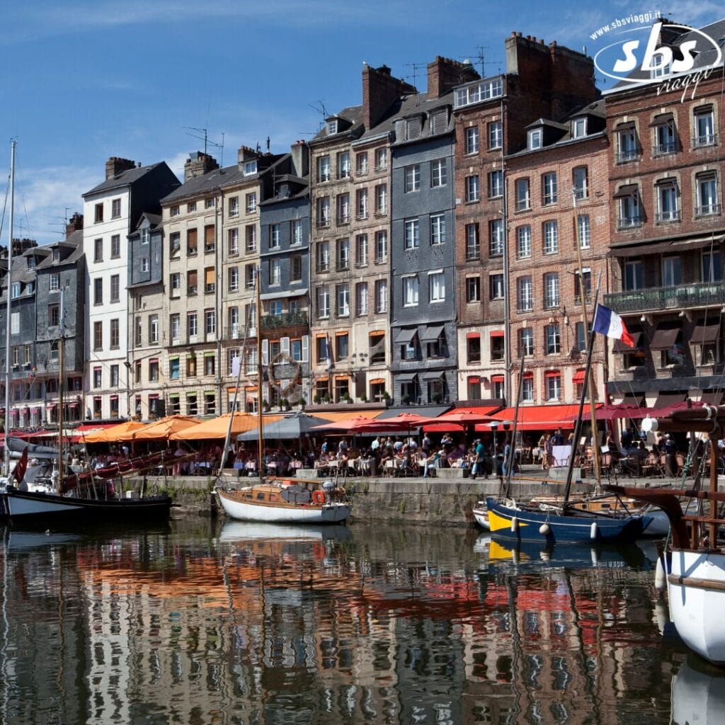 Bozza automatica: edifici storici vivaci costeggiano con grazia un lungomare, dove le barche attraccate ondeggiano dolcemente. Una bandiera francese sventola orgogliosamente in primo piano sotto un cielo azzurro e terso, completando questa scena pittoresca.