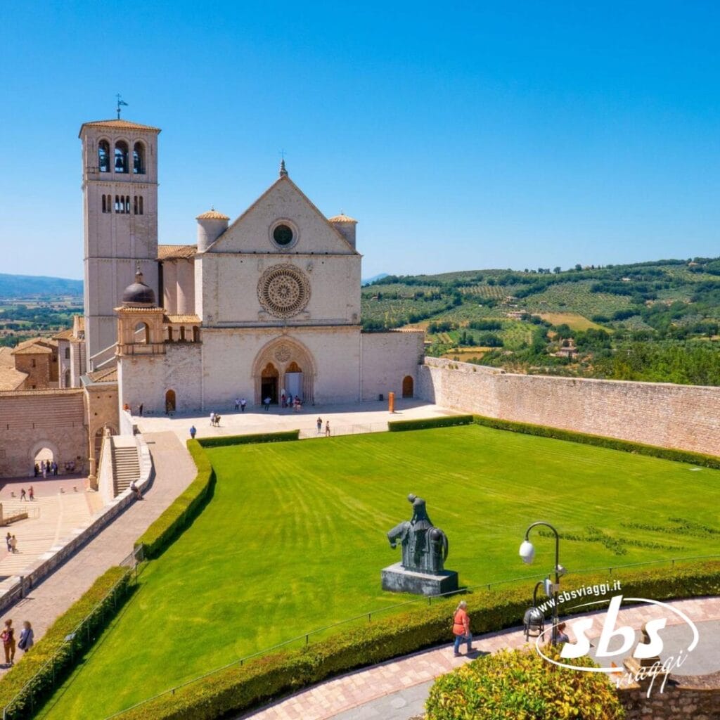 Una chiesa storica in pietra con un alto campanile si erge accanto a un prato ben curato e sentieri, emanando un fascino medievale. Una scultura e una lampada sono visibili vicino al prato, sullo sfondo di un ampio paesaggio verde che ricorda la bellezza dell'Umbria.