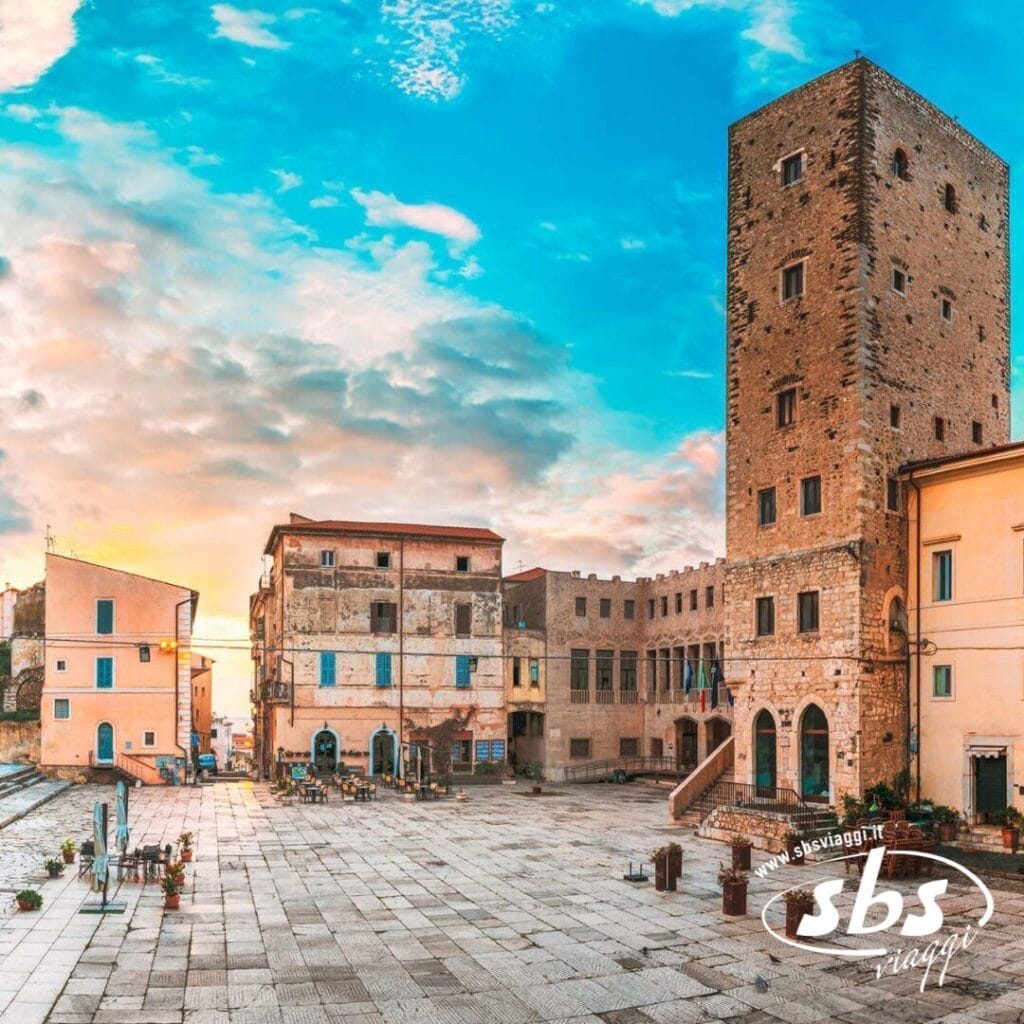 Storica piazza in pietra con torre alta, edifici circostanti e una piazza aperta e lastricata al tramonto. Piccole piante costeggiano i bordi, evocando il fascino di Ninfa e Sermoneta.
