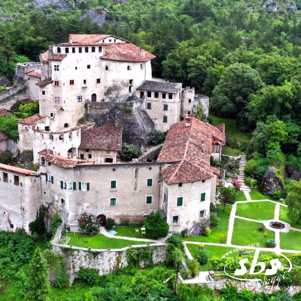 Vista aerea di edifici storici in pietra con tetti rossi, immersi nel paesaggio lussureggiante del Trentino Alto Adige, con sentieri tortuosi e giardini curati. Perfetto per un tour di Castelli. Logo nell'angolo.