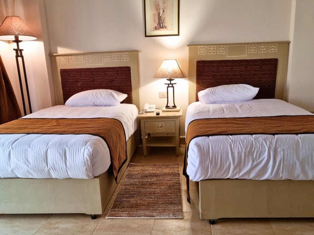 Due letti singoli con testiere marroni e coperte abbinate sono separati da un comodino con lampada. Una lampada da terra Bozza automatica e un quadro incorniciato aggiungono carattere alla stanza.