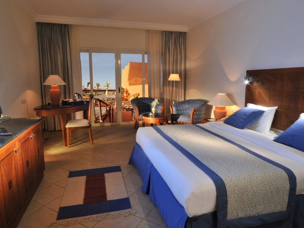 Provate la sistemazione di lusso al JAZ GRAND MARSA. Questa camera d'albergo a 5 stelle dispone di un letto matrimoniale con accenti blu, una scrivania, sedie e un tavolino. Godetevi la calda illuminazione delle lampade e la splendida vista del balcone attraverso una grande finestra.