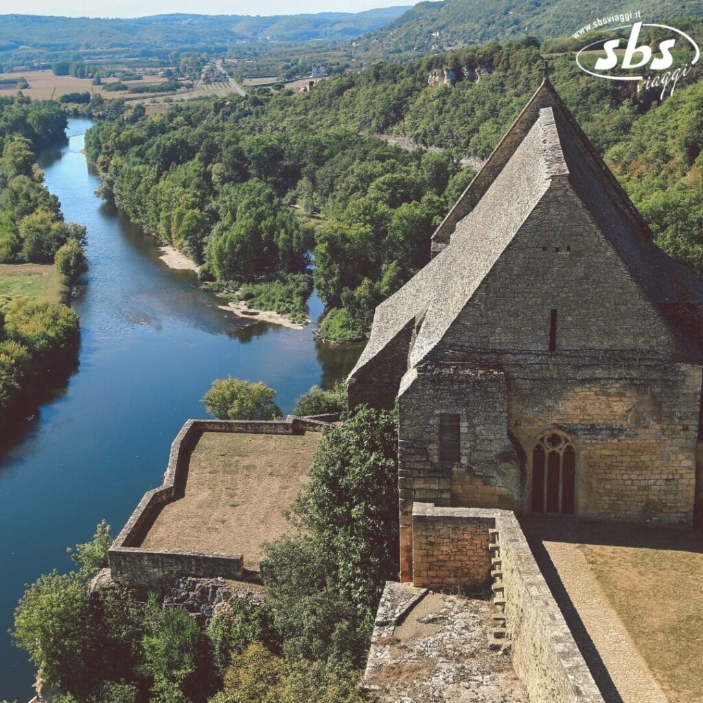 Un edificio in pietra con un tetto ripido si affaccia su un fiume e su un paesaggio lussureggiante, incarnando l'essenza della bozza automatica. Un logo con testo appare nell'angolo in alto a destra.