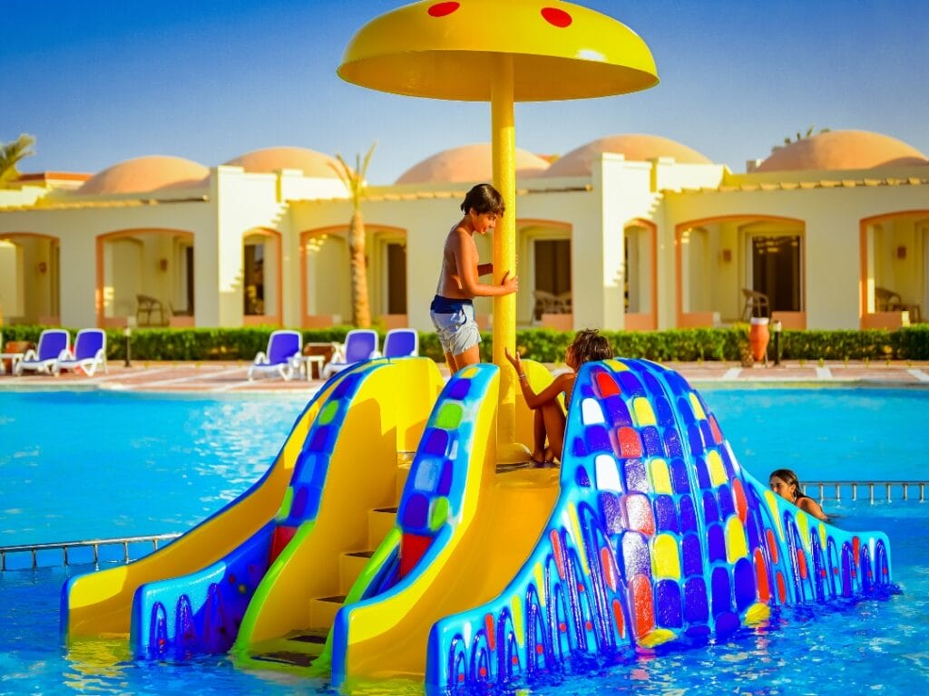 Bambini che sguazzano gioiosamente attorno a uno scivolo vivace a forma di fungo nella piscina, incorniciato da edifici e invitanti sedie a sdraio sotto il cielo azzurro brillante. La scena esplode di energia vivace e cattura una spensierata giornata estiva.