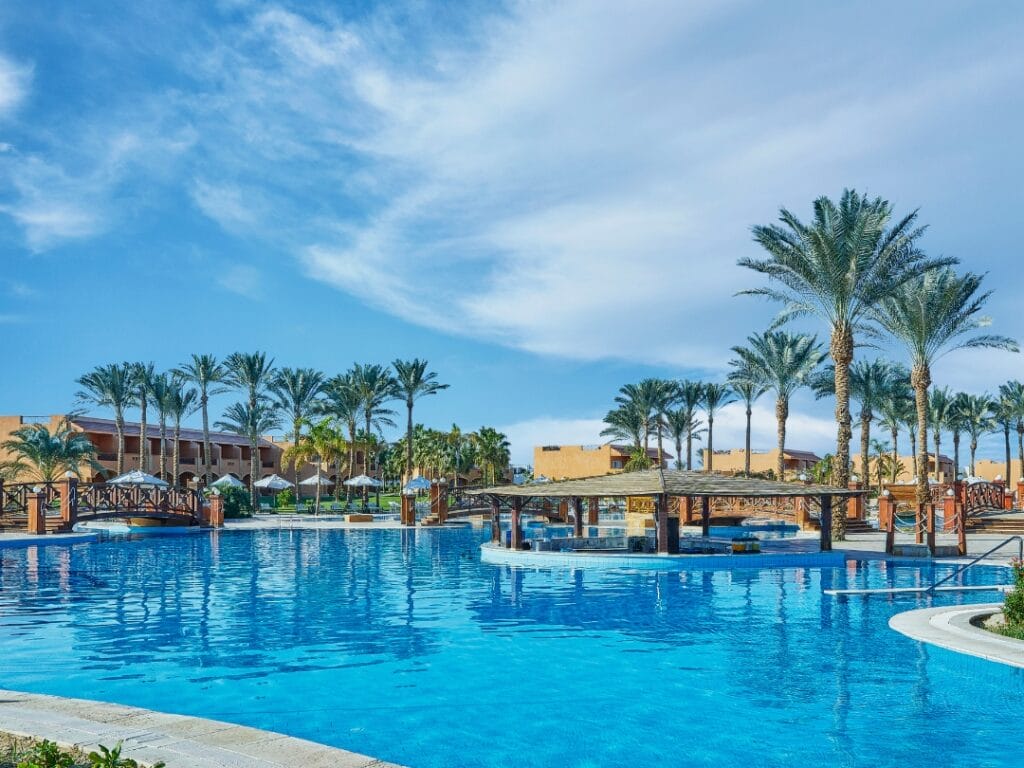 Provate il lusso del JAZ GRAND MARSA, un hotel a 5 stelle con piscina all'aperto circondata da palme ondeggianti, un incantevole ponte e un bar a bordo piscina sotto il limpido cielo azzurro.