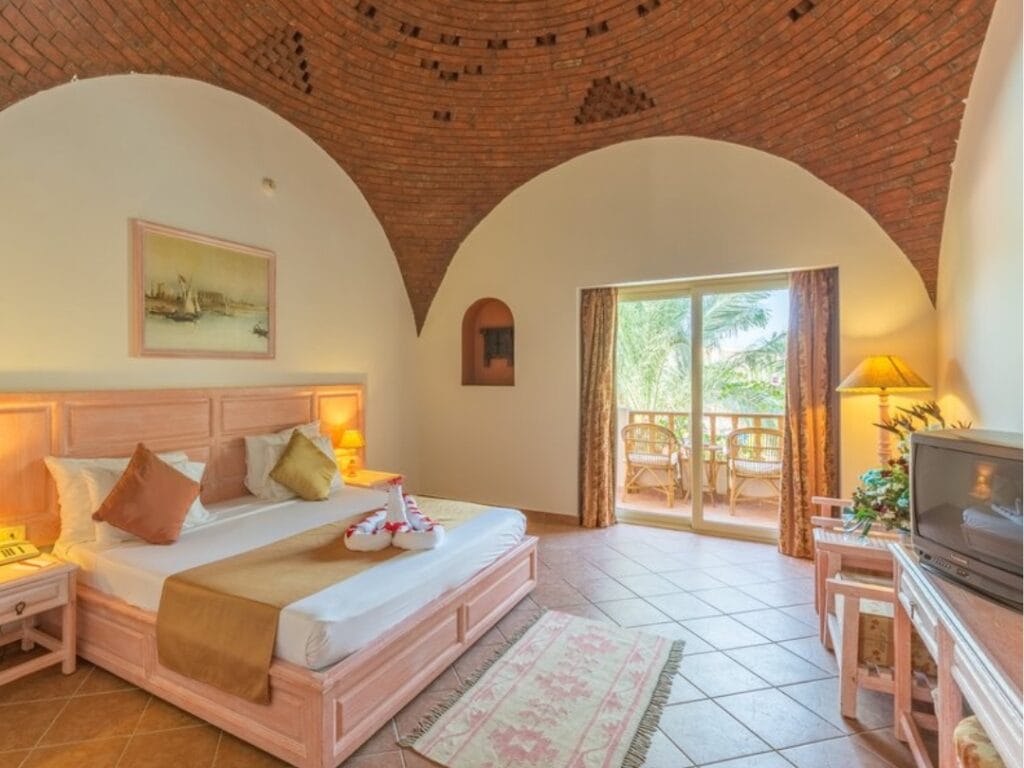 La camera d'albergo è dotata di letto, mobili in legno, TV e un incantevole soffitto a cupola in mattoni. Una porta scorrevole in vetro si apre su un balcone con palme ondeggianti all'esterno. Illuminazione calda e un asciugamano decorativo sul letto completano questa pittoresca ambientazione da bozza automatica.