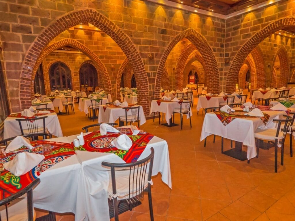 Un grande ristorante con arcate in mattoni presenta tavoli apparecchiati con tovaglie bianche e runner con motivi colorati, circondati da sedie in legno. Una calda illuminazione ambientale mette in risalto l'architettura ad arco, creando un'atmosfera invitante che ricorda il design di una bozza automatica.