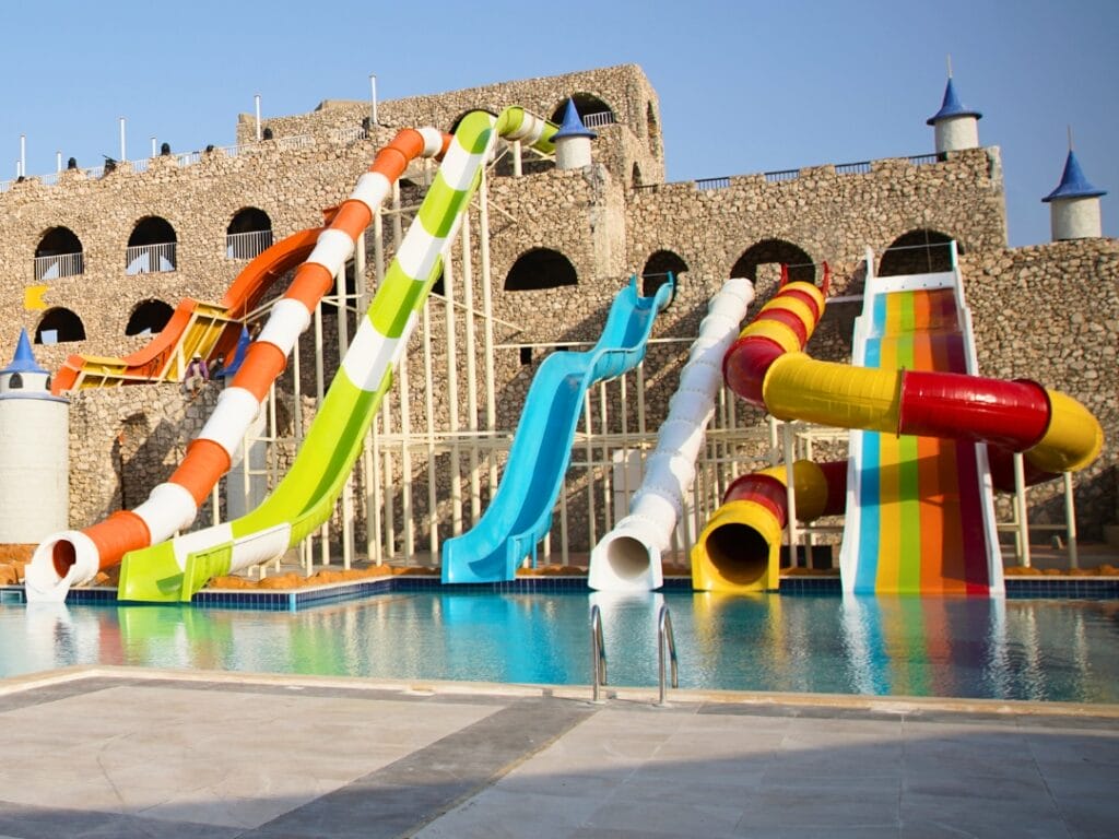 Una serie di colorati scivoli d'acqua che si riversano in una piscina si abbinano con eleganza alle strutture a torre dell'edificio in pietra sullo sfondo, il tutto sotto un limpido cielo azzurro, in un mix perfetto di divertimento e architettura.
