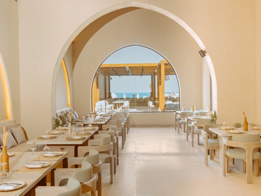 Una sala da pranzo con tavoli e sedie ordinatamente disposti, tutti in una combinazione di colori beige, si trova all'interno del lusso del Sunrise Anjum Resort. Le grandi aperture ad arco offrono una vista mozzafiato su una terrazza esterna e sul cielo azzurro oltre.