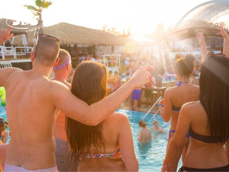 Persone in costume da bagno si divertono a una vivace festa in piscina a tema Vacanza, con alcuni che ballano e altri in acqua, sotto un cielo soleggiato.
