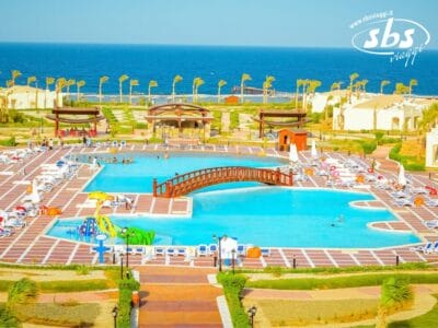 L'area della piscina del resort, dotata di un ponte di legno e circondata da sedie a sdraio, palme ed erba, si affaccia sull'oceano: un perfetto esempio di serena bellezza costiera.