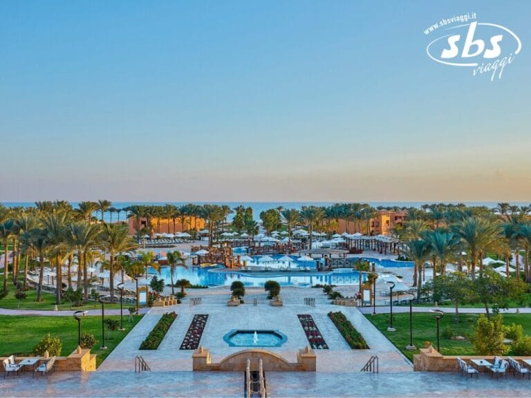 Vista del resort con più piscine, palme e vegetazione circostante sotto un cielo azzurro terso. Logo nell'angolo in alto a destra.