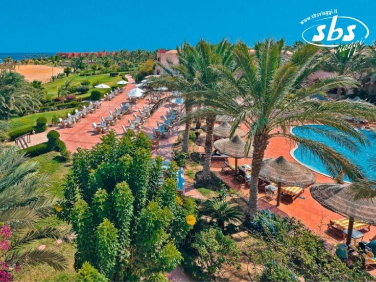 Vista aerea di un resort con piscina, lettini prendisole, palme e giardini paesaggistici. L'oceano è visibile sullo sfondo sotto un cielo azzurro terso, catturando un momento perfetto che sembra realizzato con la precisione di una bozza automatica.