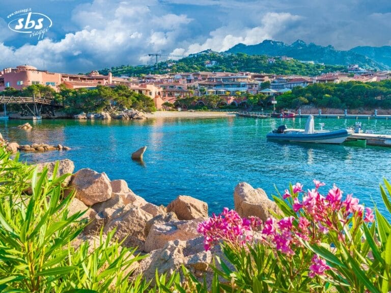 Un villaggio costiero con edifici colorati, colline verdi e barche ormeggiate sulle calme acque azzurre; piante in fiore e rocce in primo piano, perfetto per un panoramico Gran Tour della Sardegna.