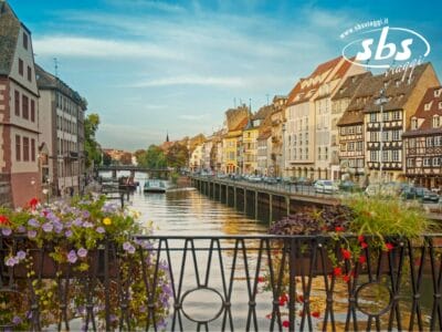 Una vista panoramica di un canale fiancheggiato da edifici colorati, barche e fiori su una ringhiera a Strasburgo, in Francia, che ricorda un grazioso dipinto di Bozza automatica.
