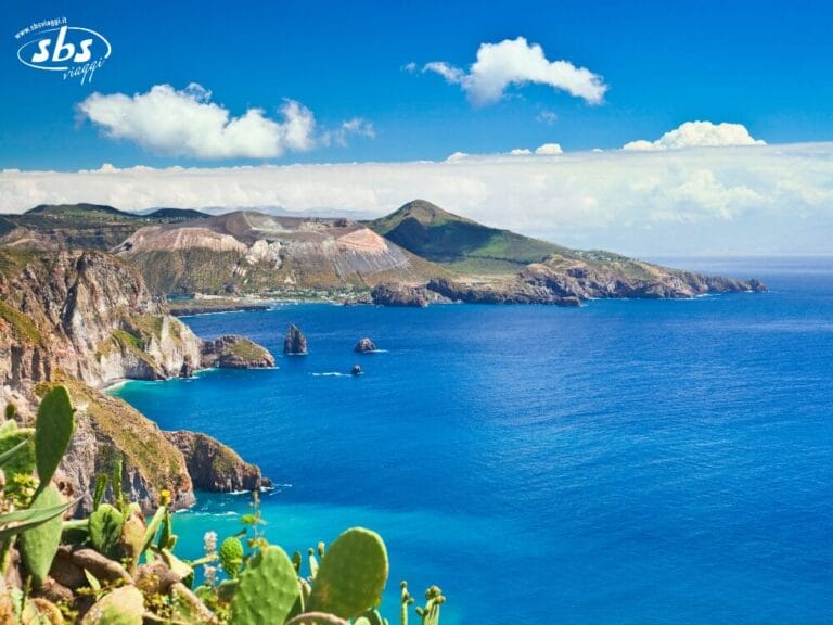 Paesaggio costiero con scogliere rocciose, verdi colline e l'oceano azzurro sotto un cielo parzialmente nuvoloso; in primo piano sono visibili piante di cactus, che evocano un senso di libertà che si può provare durante un tour delle Isole Eolie.