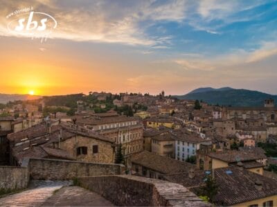 La vista del tramonto sulla città storica dell'Umbria mette in mostra edifici in pietra incastonati tra colline medievali. Il cielo dipinge una tela parzialmente nuvolosa con una splendida sfumatura arancione e blu, creando un'atmosfera incantevole.