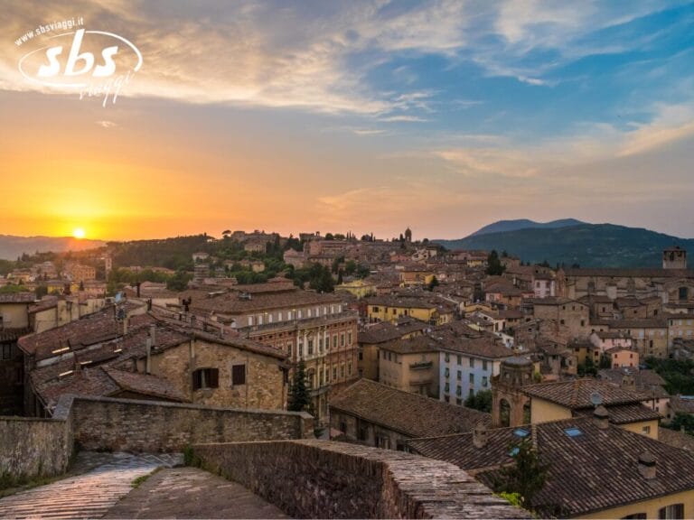 La vista del tramonto sulla città storica dell'Umbria mette in mostra edifici in pietra incastonati tra colline medievali. Il cielo dipinge una tela parzialmente nuvolosa con una splendida sfumatura arancione e blu, creando un'atmosfera incantevole.
