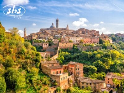 Una vista panoramica di una storica cittadina italiana nel cuore di Siena, con edifici in terracotta e colline verdeggianti sotto un cielo azzurro, che riecheggia gli affascinanti paesaggi della Val d'Orcia.