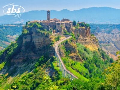 Villaggio in cima a una collina con edifici in pietra e una torre dell'orologio, collegato all'area circostante da uno stretto ponte pedonale. Verde lussureggiante, montagne sullo sfondo sotto un cielo limpido: scopri il fascino di Etrusco qui nel sorriso dell'Etrusco.