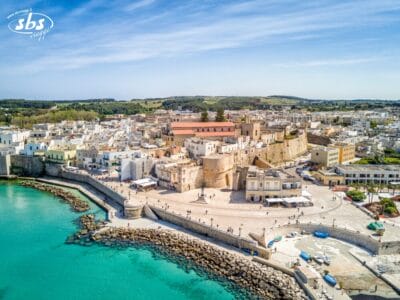 Vista aerea di una cittadina costiera del Salento, con edifici storici in pietra, un lungomare e un mare turchese, circondata da case bianche e verde: uno dei veri tesori del Salento.