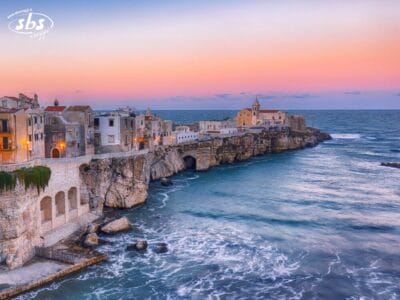 Una città costiera italiana arroccata su aspre scogliere al tramonto, con edifici storici che si affacciano sul mare calmo e azzurro e sulle dolci onde sottostanti, evoca il fascino della spettacolare costa del Gargano.