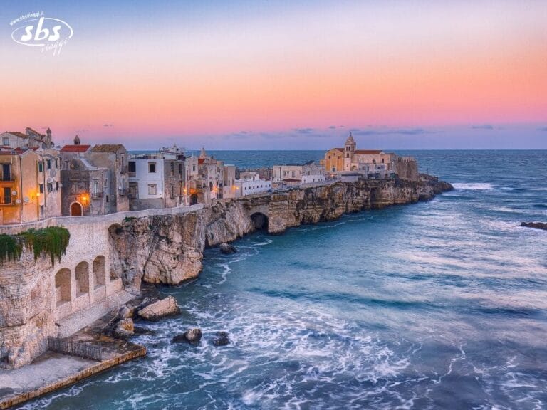 Una città costiera italiana arroccata su aspre scogliere al tramonto, con edifici storici che si affacciano sul mare calmo e azzurro e sulle dolci onde sottostanti, evoca il fascino della spettacolare costa del Gargano.
