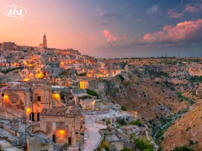 Gli antichi edifici in pietra di Matera, in Italia, sono illuminati al tramonto e il loro caldo chiarore mette in risalto il paesaggio roccioso e le colline lontane sotto un cielo colorato: uno scenario incantevole nel cuore della Città Bianche.