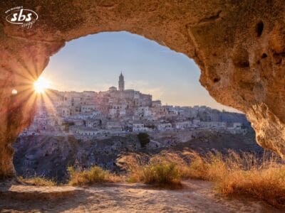 Una veduta dell'antica città collinare di Matera al tramonto, ripresa attraverso l'apertura di una grotta rocciosa, con i raggi del sole che brillano attraverso: un momento perfetto di un'avventura del Gran Tour Puglia.