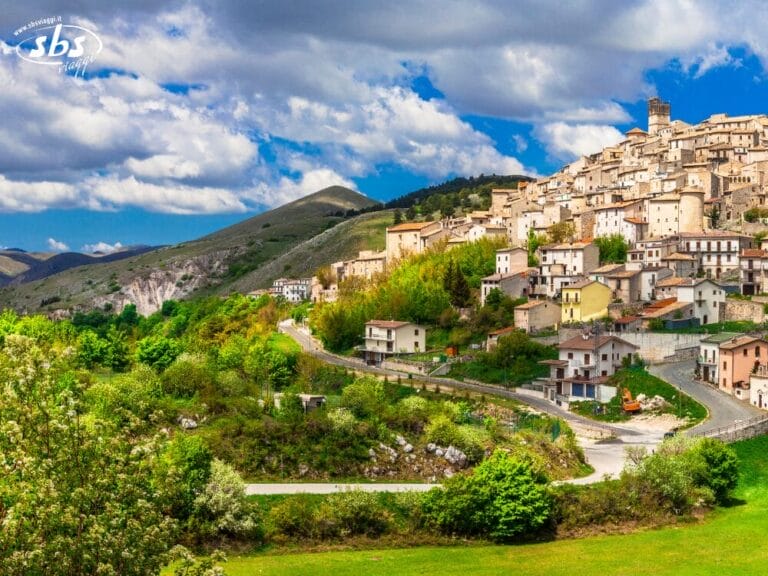Un borgo collinare con case in pietra e una torre, circondato da alberi verdi e montagne sotto un cielo parzialmente nuvoloso: uno scenario perfetto da scoprire durante un tour in Abruzzo.