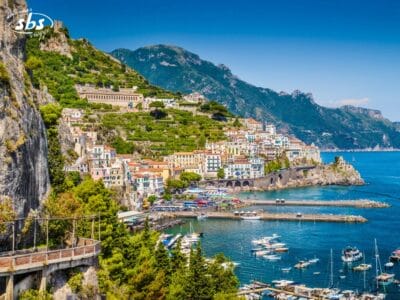 Colorata cittadina costiera su ripide colline che dominano una baia azzurra, con barche ormeggiate in un porto turistico e circondata da montagne verdi: un'incantevole perla della Costiera Amalfitana.