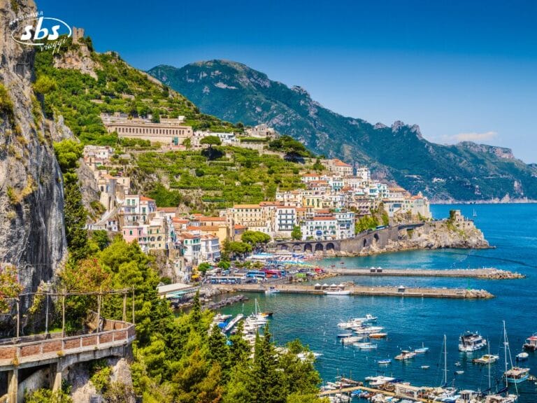 Colorata cittadina costiera su ripide colline che dominano una baia azzurra, con barche ormeggiate in un porto turistico e circondata da montagne verdi: un'incantevole perla della Costiera Amalfitana.