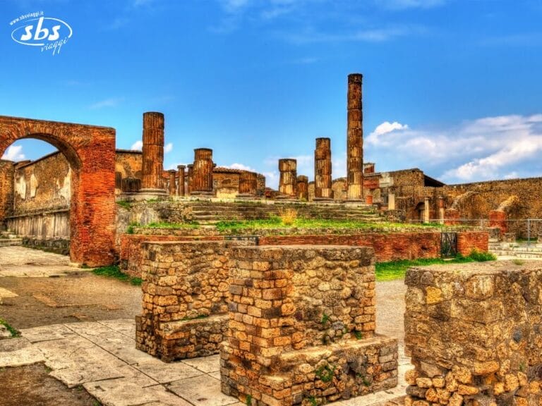 Rovine di antiche strutture in pietra e mattoni con colonne e un arco sotto un cielo azzurro, probabilmente parte di un sito archeologico storico nei pressi di Napoli.
